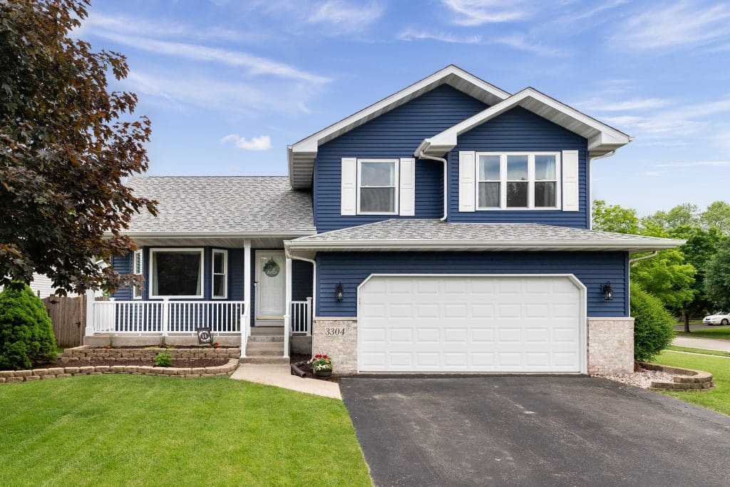 Exterior of a suburban home with rain gutters, blue siding, a white front porch, white shutters and landscaping.